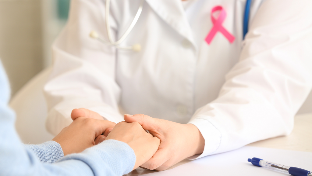 Médica com jaleco branco e fita rosa da campanha outubro rosa segurando as mãos de uma paciente com suéter azul